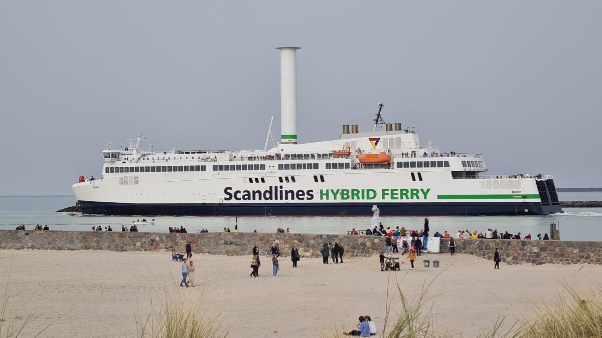 Blick vom Strand auf eine Scandlines-Fähre mit dem großen Flettner-Rotor in Schornsteinform