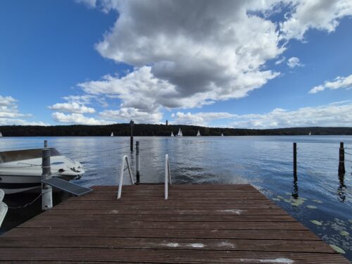 blick auf einen steg mit einer leiter, im wasser ein paar segelbooten, am anderen ufer sieht man wald.